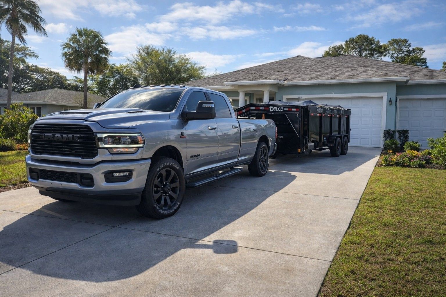 Black dumpster on a driveway in front of a white garage door; a house and tree are in the background.