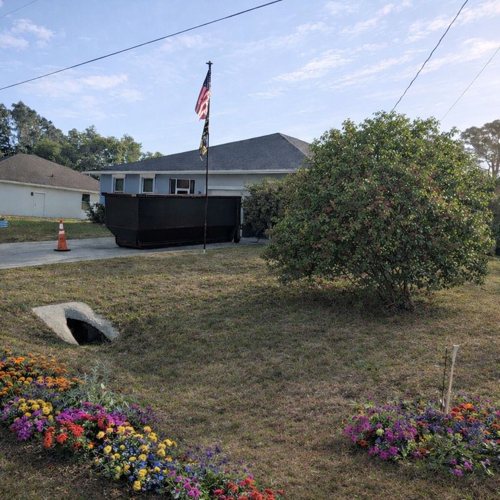 House with American flag, dumpster in driveway, tree and flowers in foreground.
