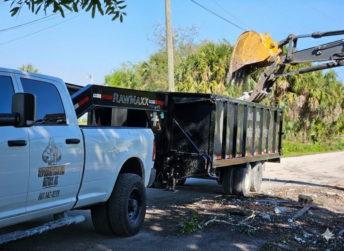 White truck towing a trailer being loaded by an excavator bucket. Bright sunny day.