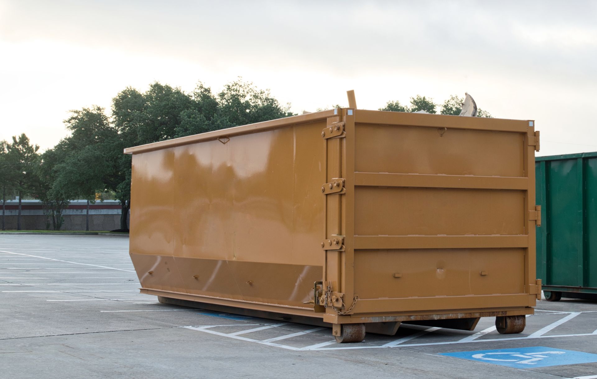 Brown dumpster in a parking lot, near green container, trees in the background.