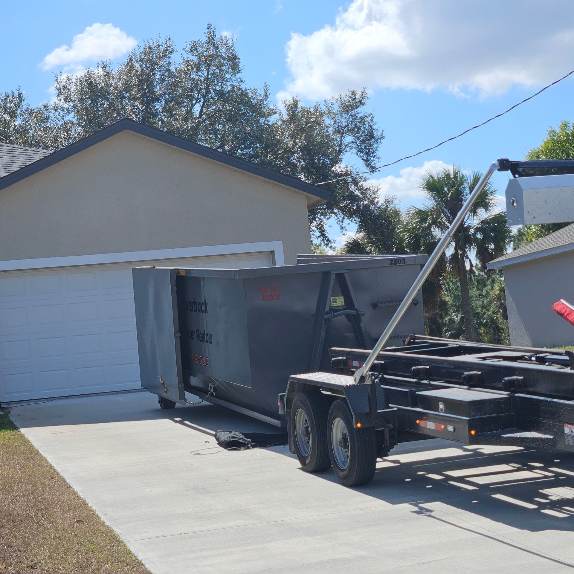 A dumpster on a trailer parked in a driveway next to a garage under a blue sky.