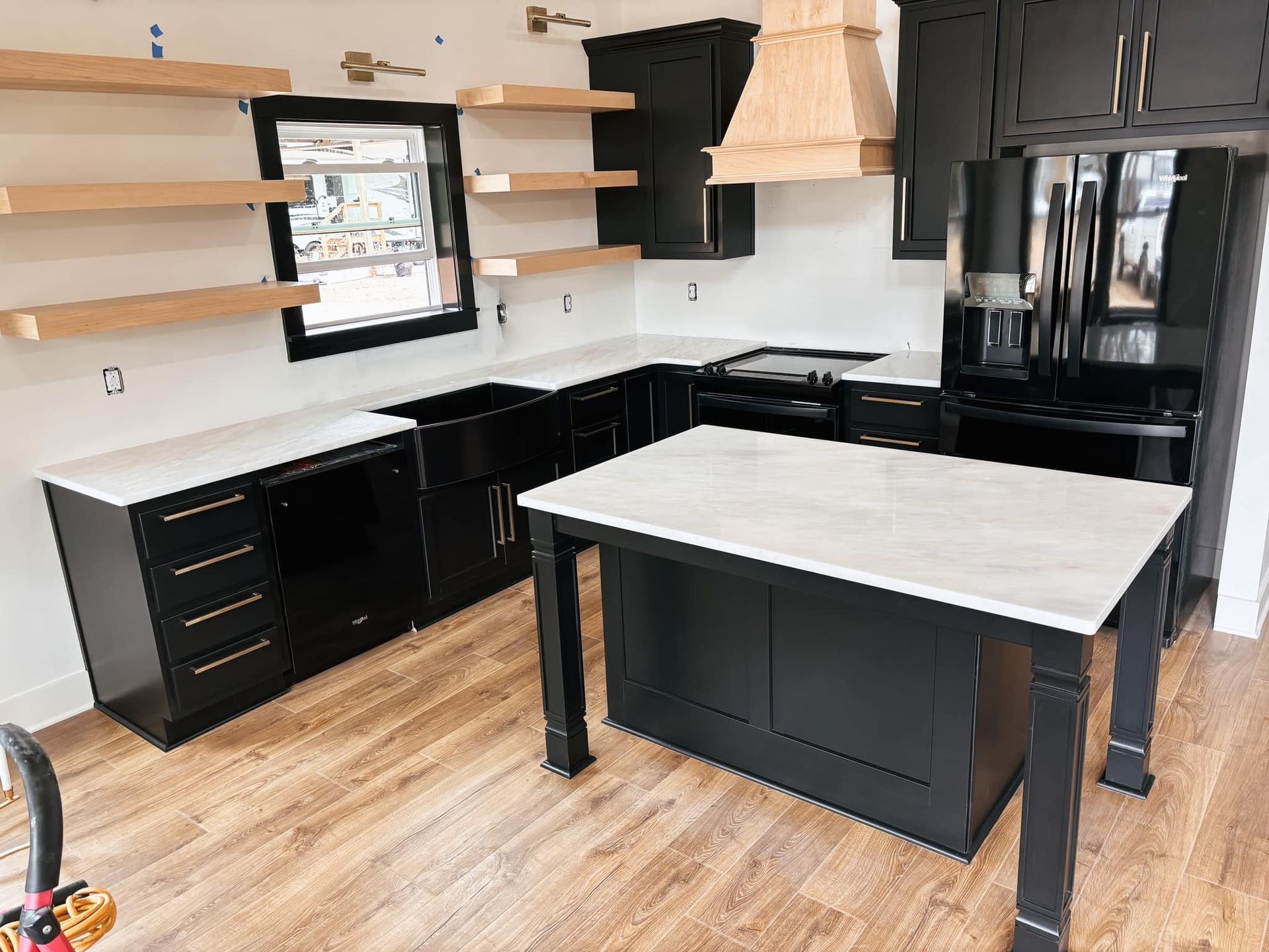 A modern kitchen featuring black cabinets, white countertops, a center island, wooden floating shelves, and a range hood.