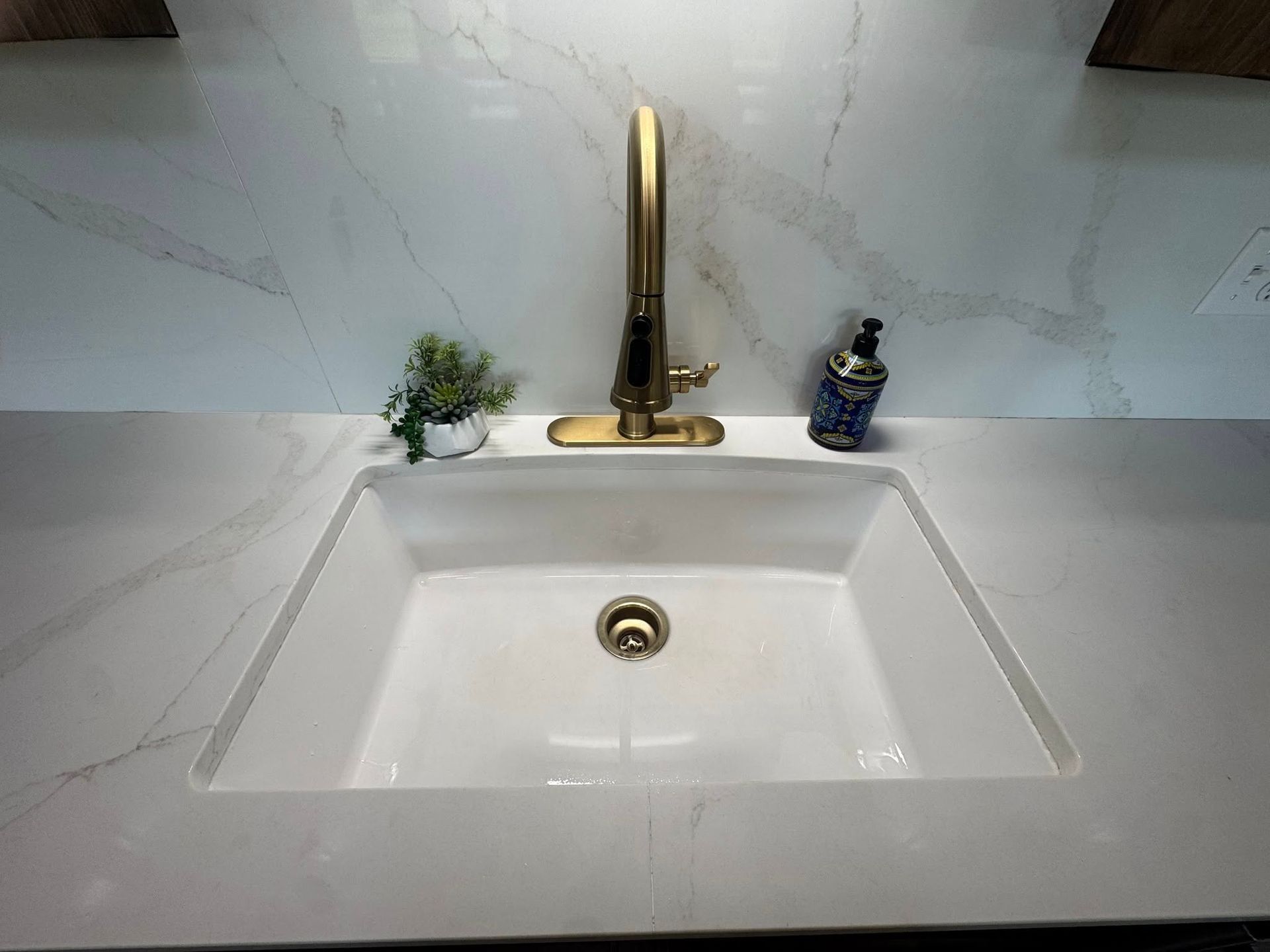 A white rectangular sink with a gold faucet, a small potted plant, and a soap dispenser on a marble-patterned counter.