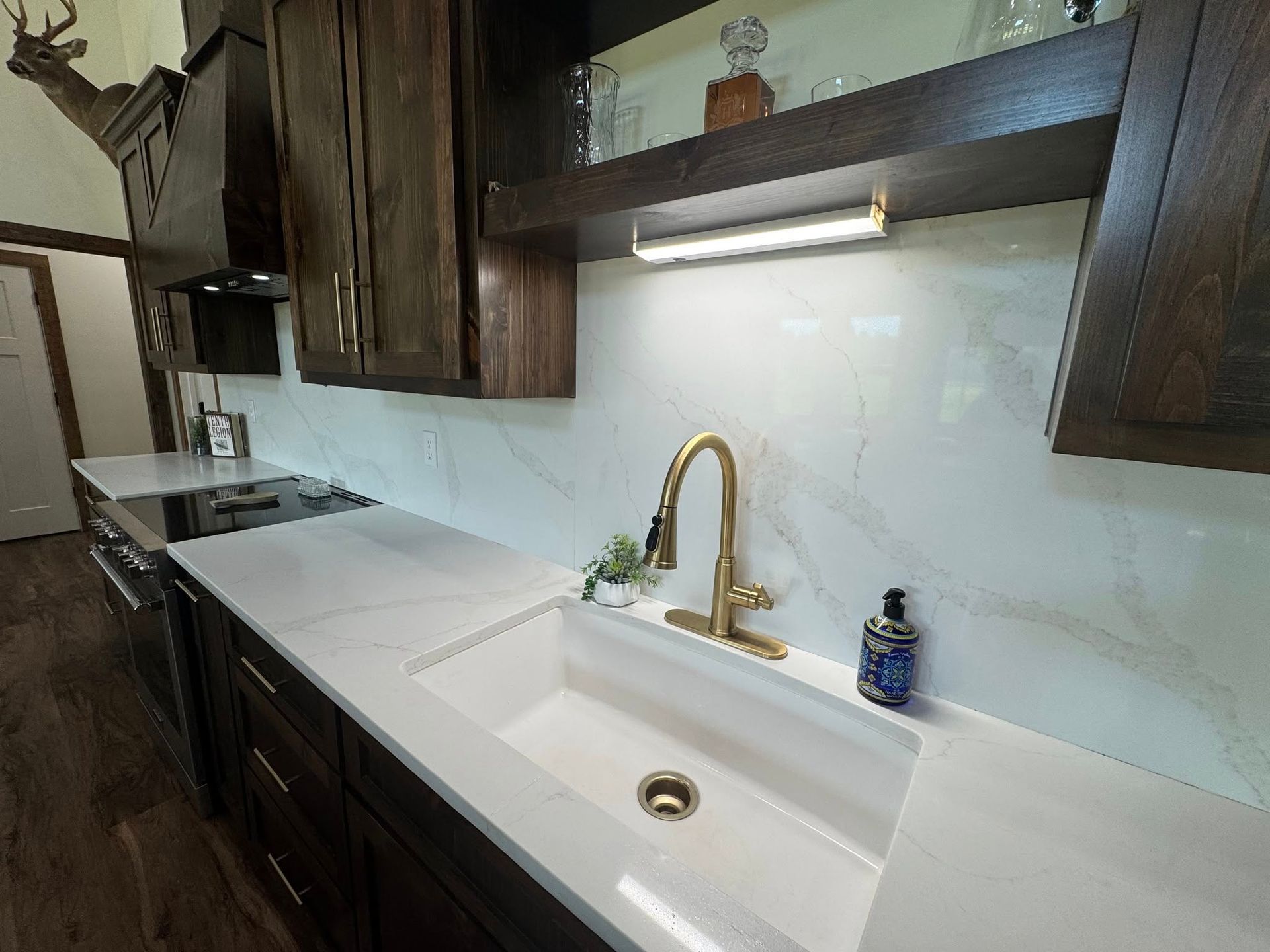 A kitchen with dark wood cabinets, white marble-style backsplash and counters, a white farmhouse sink, and gold faucet.