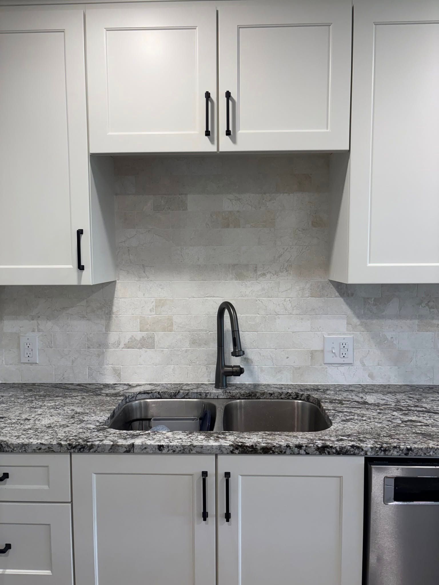 A kitchen sink area featuring a granite countertop, light stone backsplash, white cabinets, and a matte black faucet.