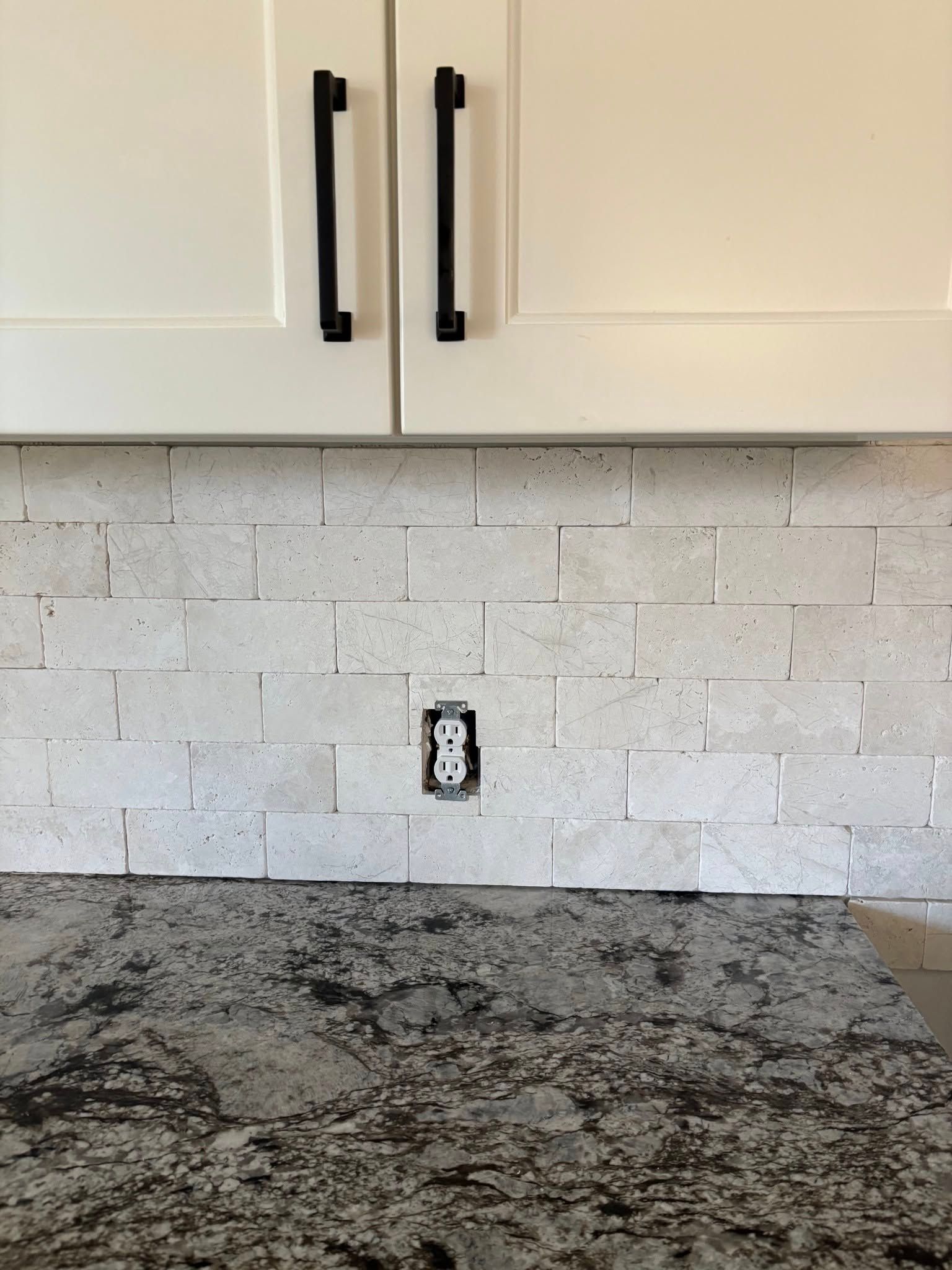 A kitchen backsplash of light-colored, textured rectangular tiles with a central electrical outlet above a granite counter.