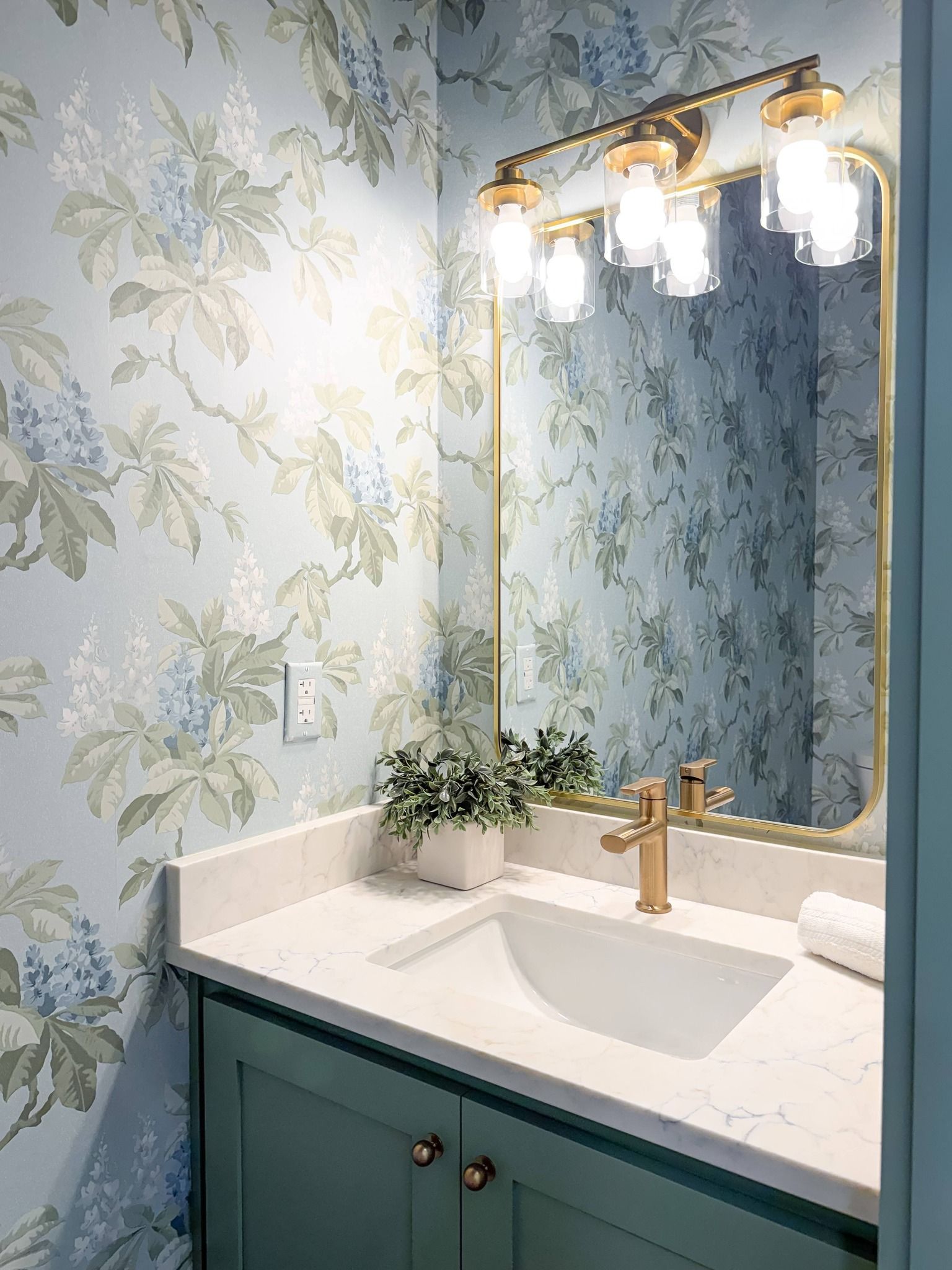 A bathroom vanity with a green cabinet, white countertop, gold faucet, and a wall mirror against light blue floral wallpaper.