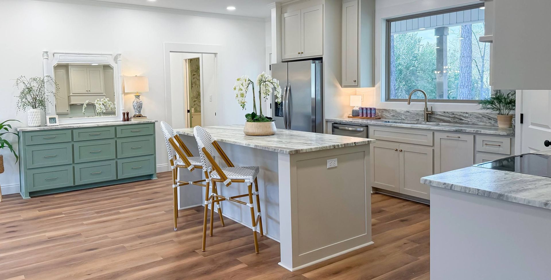 A kitchen featuring a light gray island with two stools, a sage green dresser against the wall, and hardwood flooring.