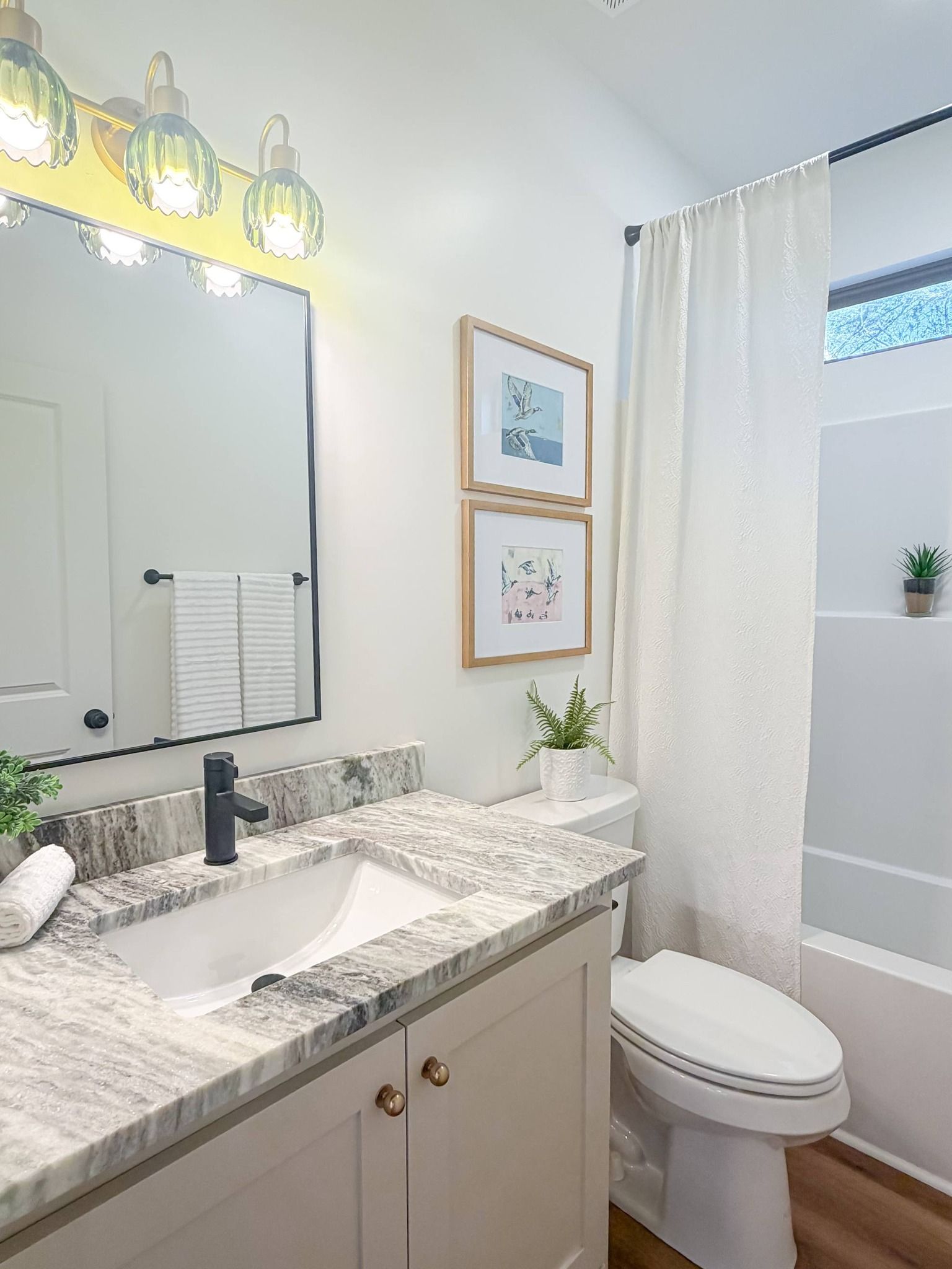 Modern bathroom with a marble vanity, rectangular sink, gold light fixture, framed wall art, and white shower curtain.