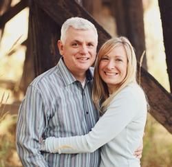 Smiling couple embracing outdoors near wooden beams, with warm sunlight in the background.