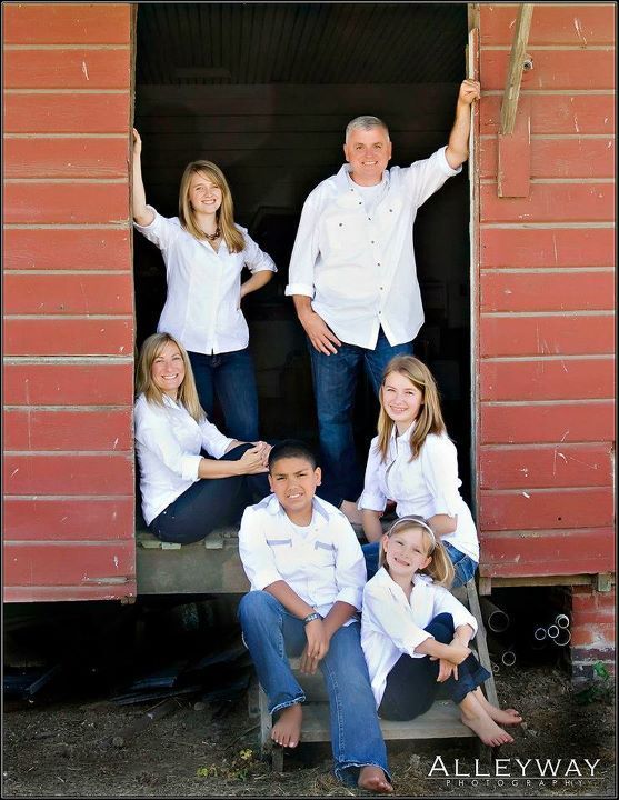 Six people in white shirts and dark pants standing barefoot against a wooden wall, posing together