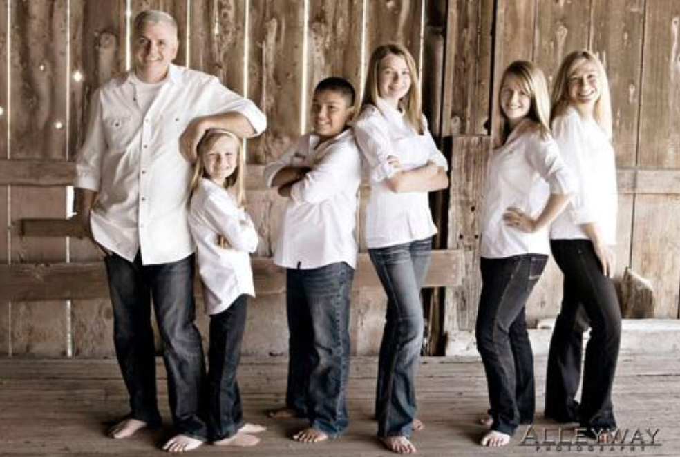 Six people in white shirts and dark pants standing barefoot against a wooden wall, posing together