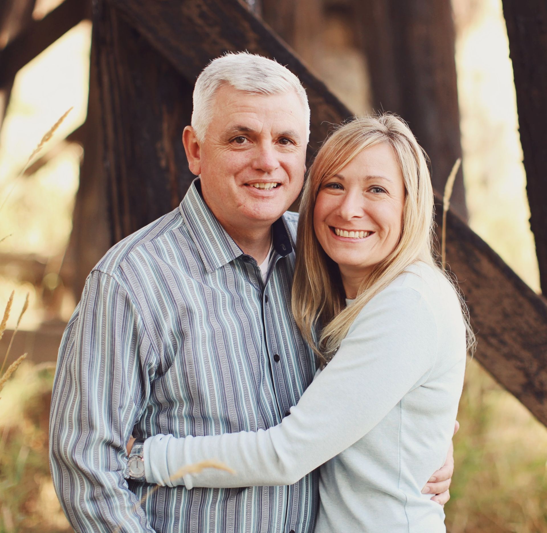 Smiling couple embracing outdoors near wooden beams, with warm sunlight in the background.