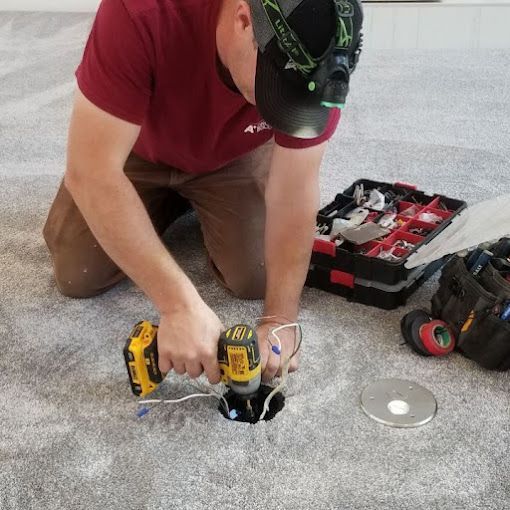 Person installing an electrical outlet in carpet using a drill; tools and components visible.