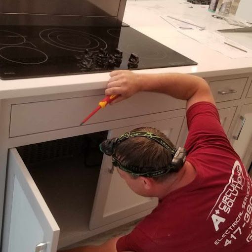 Electrician working on electrical components under a kitchen countertop with a stove.