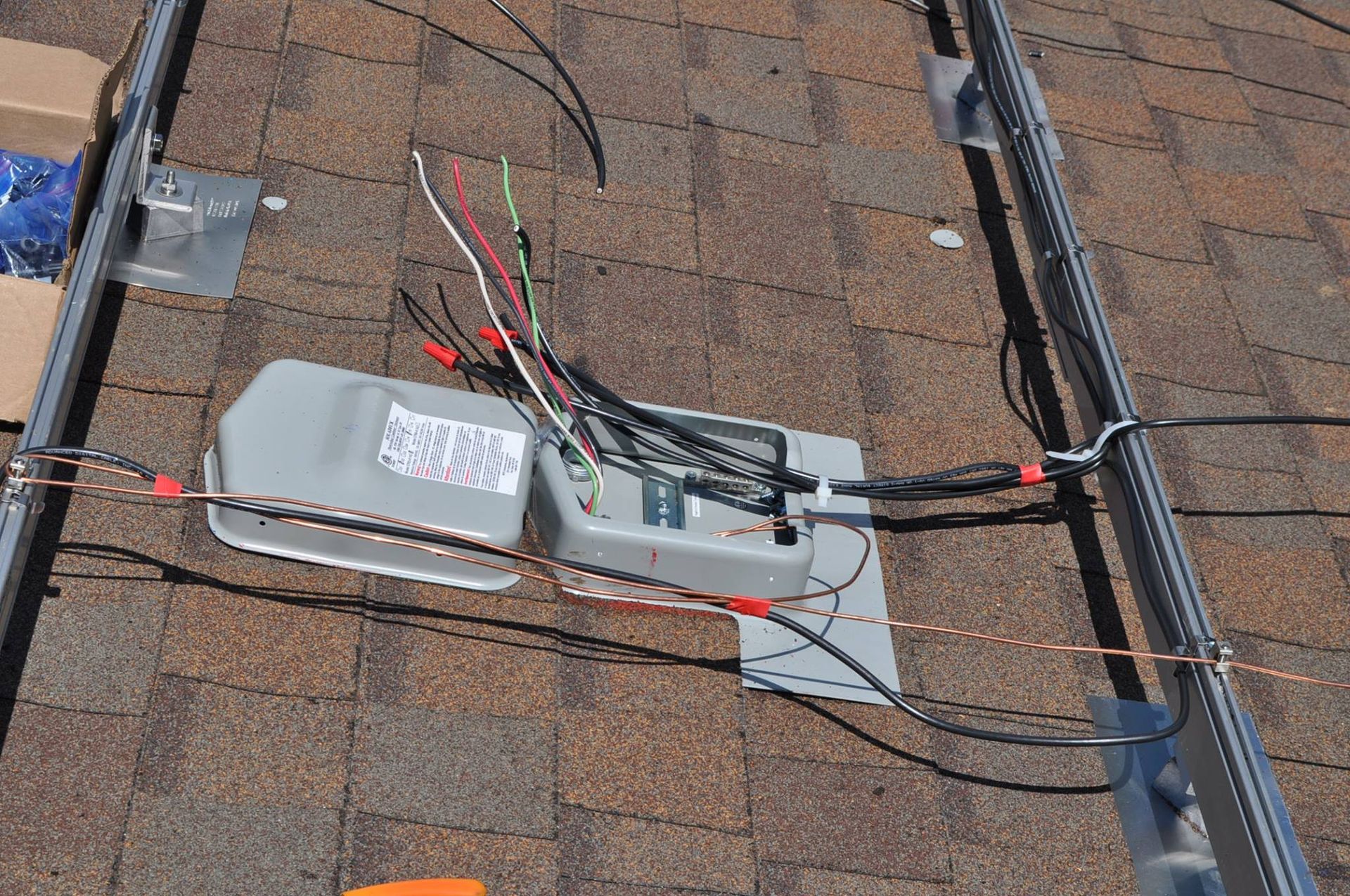 Solar panel wiring on a brown shingled roof, with gray electrical box and metal support rails.