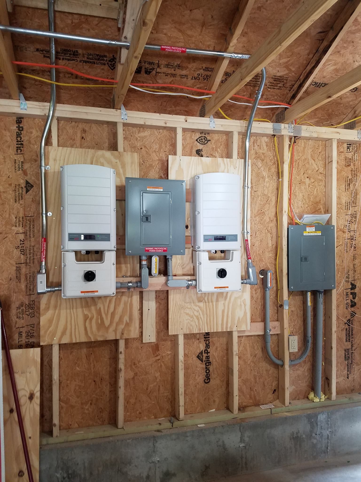 Solar panel inverters mounted on plywood in a building, with electrical conduit and wiring.