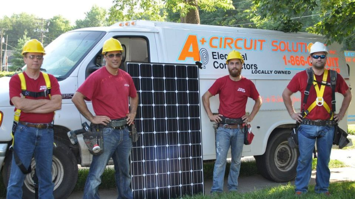 Four workers in hard hats stand with a solar panel and a company van that says 