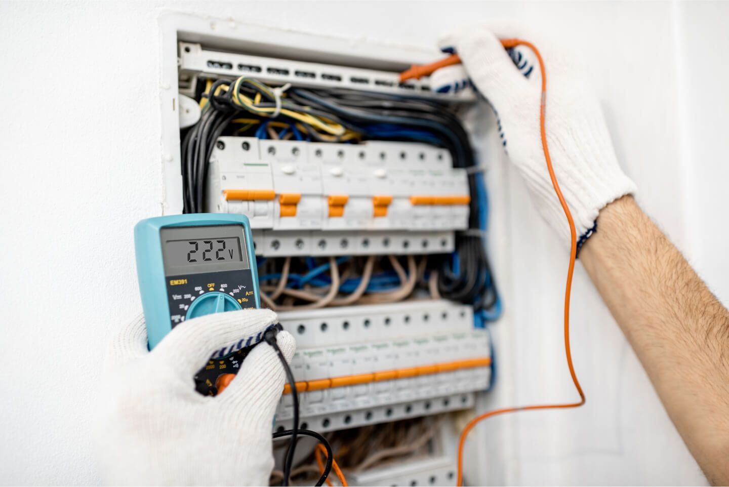 Electrician using a multimeter to test a circuit breaker panel. Hands in white gloves. Digital display reads 222.