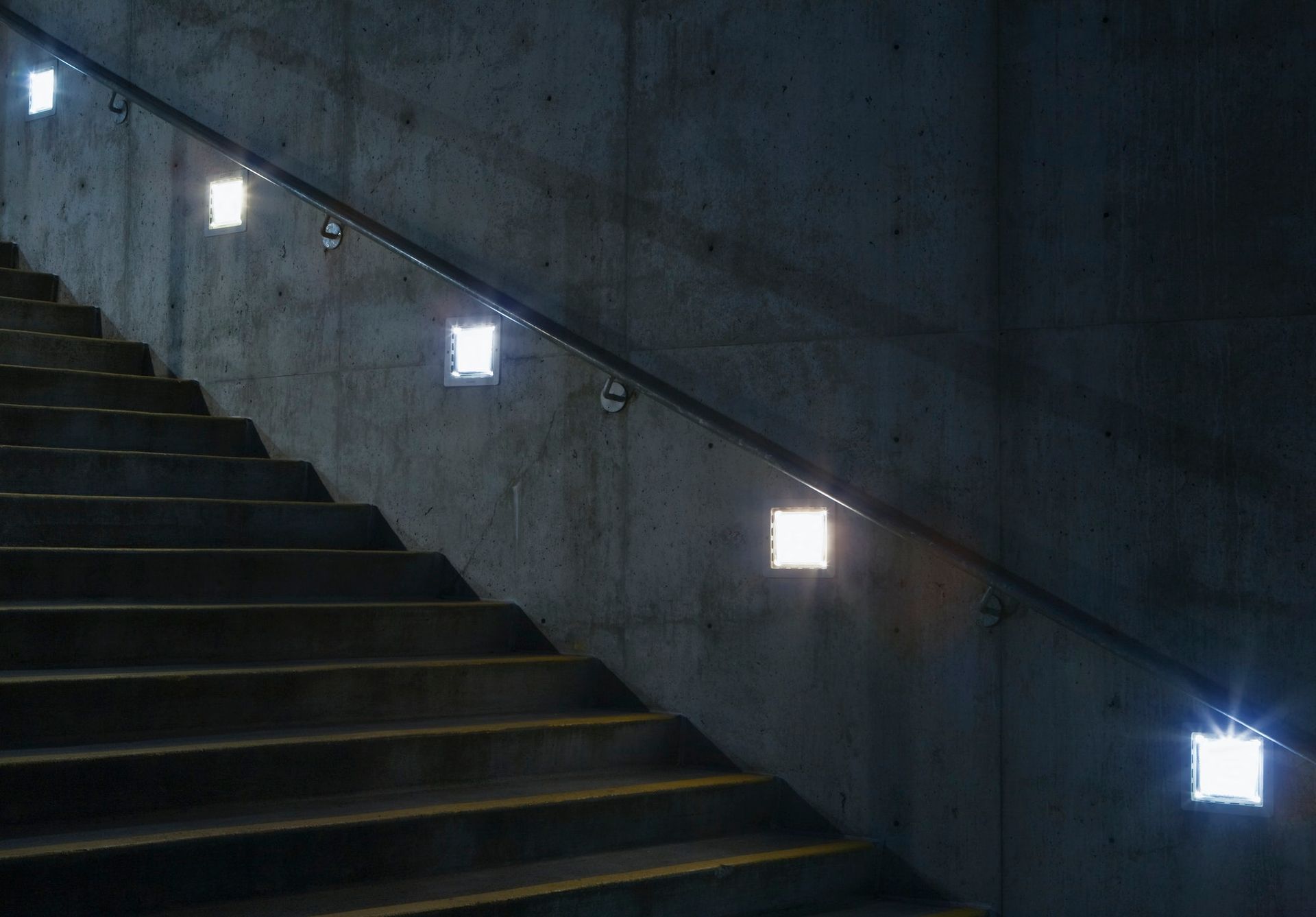 Concrete staircase with built-in square lights along the wall and handrail, illuminated steps and wall in shadows.