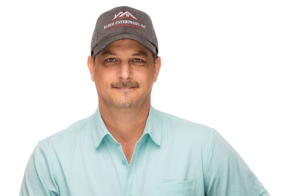 A man wearing a hat and a light blue shirt is standing in front of a white background.