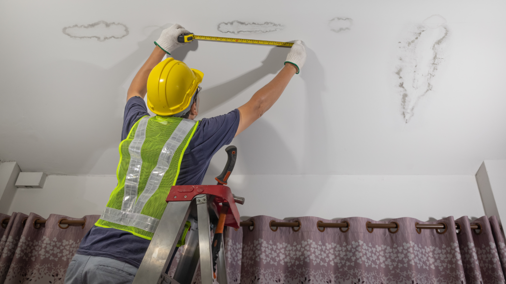 A man is measuring a wall with a tape measure while standing on a ladder.