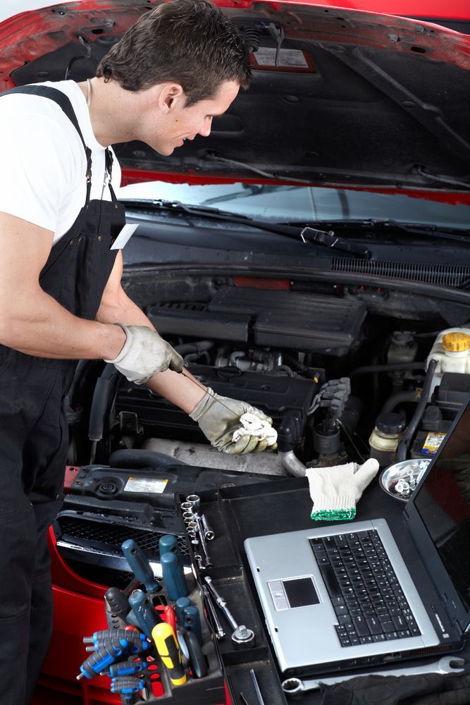 Mechanic in coveralls inspecting car engine, gloves on. Laptop and tools visible — Drinnan's Diesel & Automotive Repairs In Gladstone Central, QLD