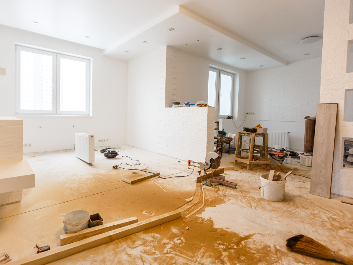 Interior of a room under renovation, white walls, scattered tools, wood debris on the floor, two windows.