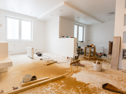 Interior of a room under renovation, white walls, scattered tools, wood debris on the floor, two windows.