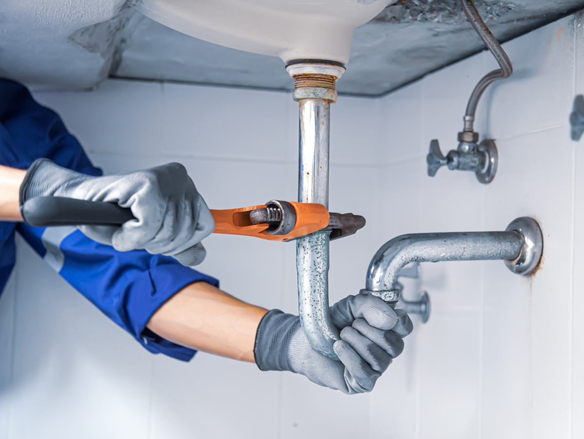 Plumber wearing gloves using a wrench on pipes under a sink.