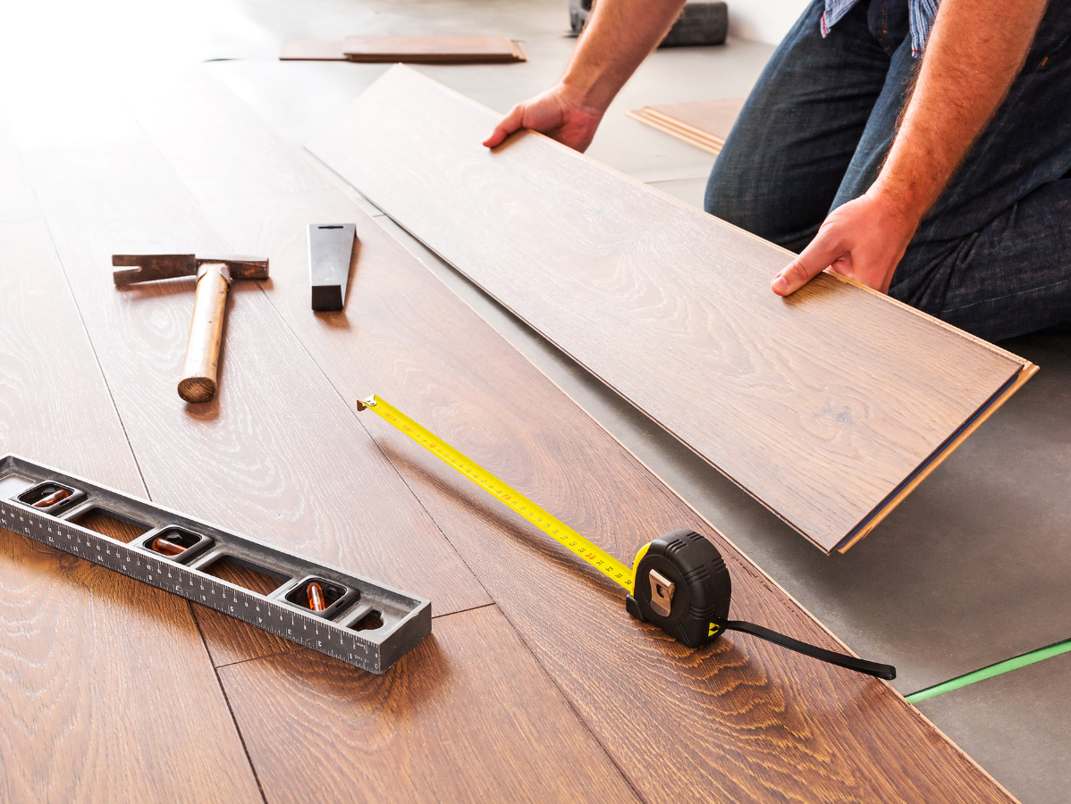 Person installing wooden flooring with tools: hammer, level, tape measure.