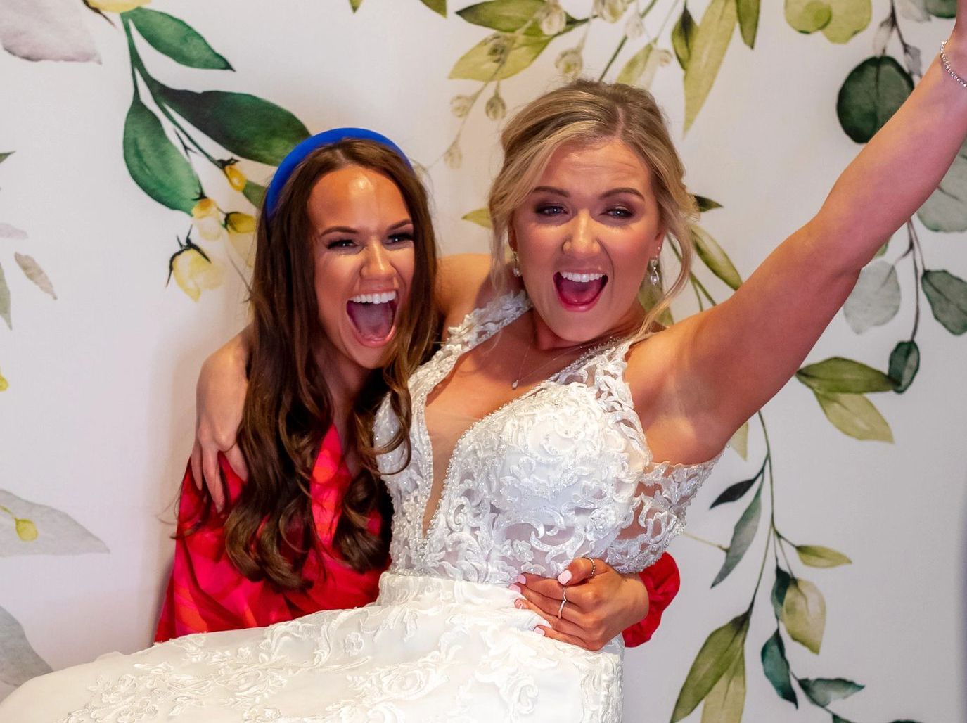 A bride and her bridesmaid are posing for a picture in front of a floral wall.