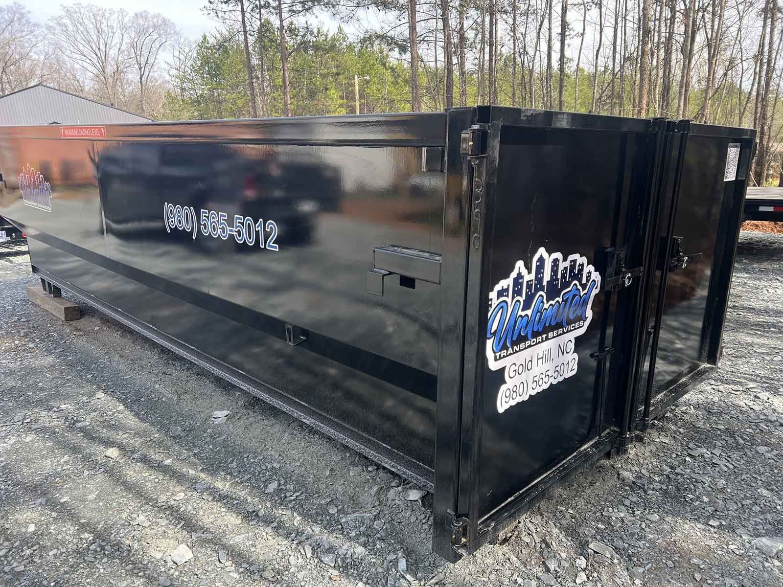 A large black dumpster is sitting on top of a dirt road.