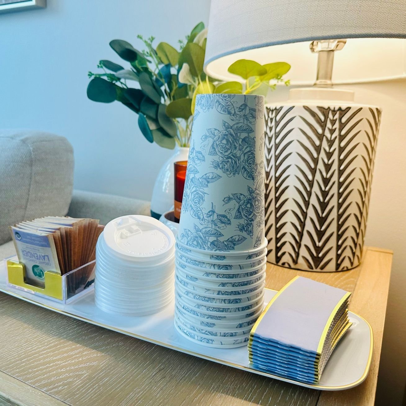 Tray with stacked cups, lids, napkins, and tea bags on a side table next to a lamp and greenery.