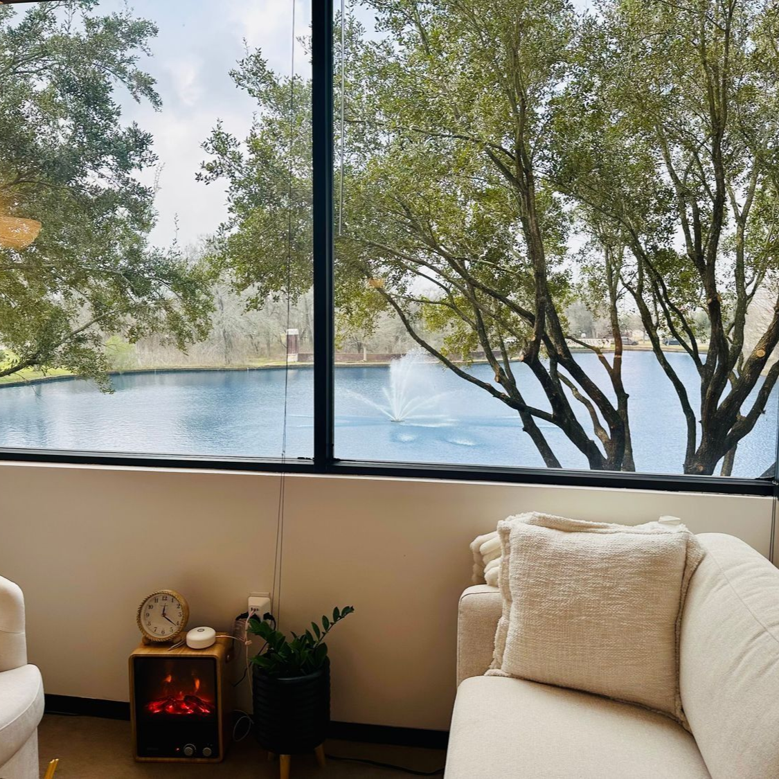 Cozy interior with a lakeside view. Cream-colored couch, small fireplace, plants, and large window with fountain in lake.