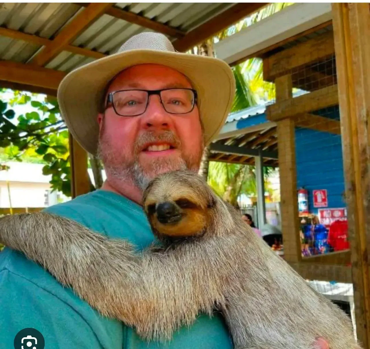 A man wearing a hat and glasses is holding a sloth