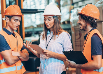 Three people in a warehouse: one woman, two men. Woman holds clipboard, gesturing. Men wear safety vests, hard hats, discussing.