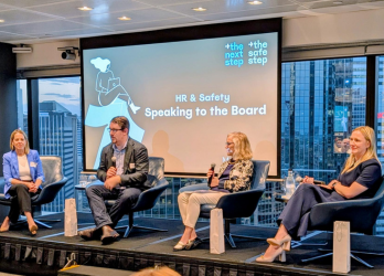 A panel of four professionals sits on chairs on a stage in front of a presentation screen titled HR & Safety: Speaking to the Board.