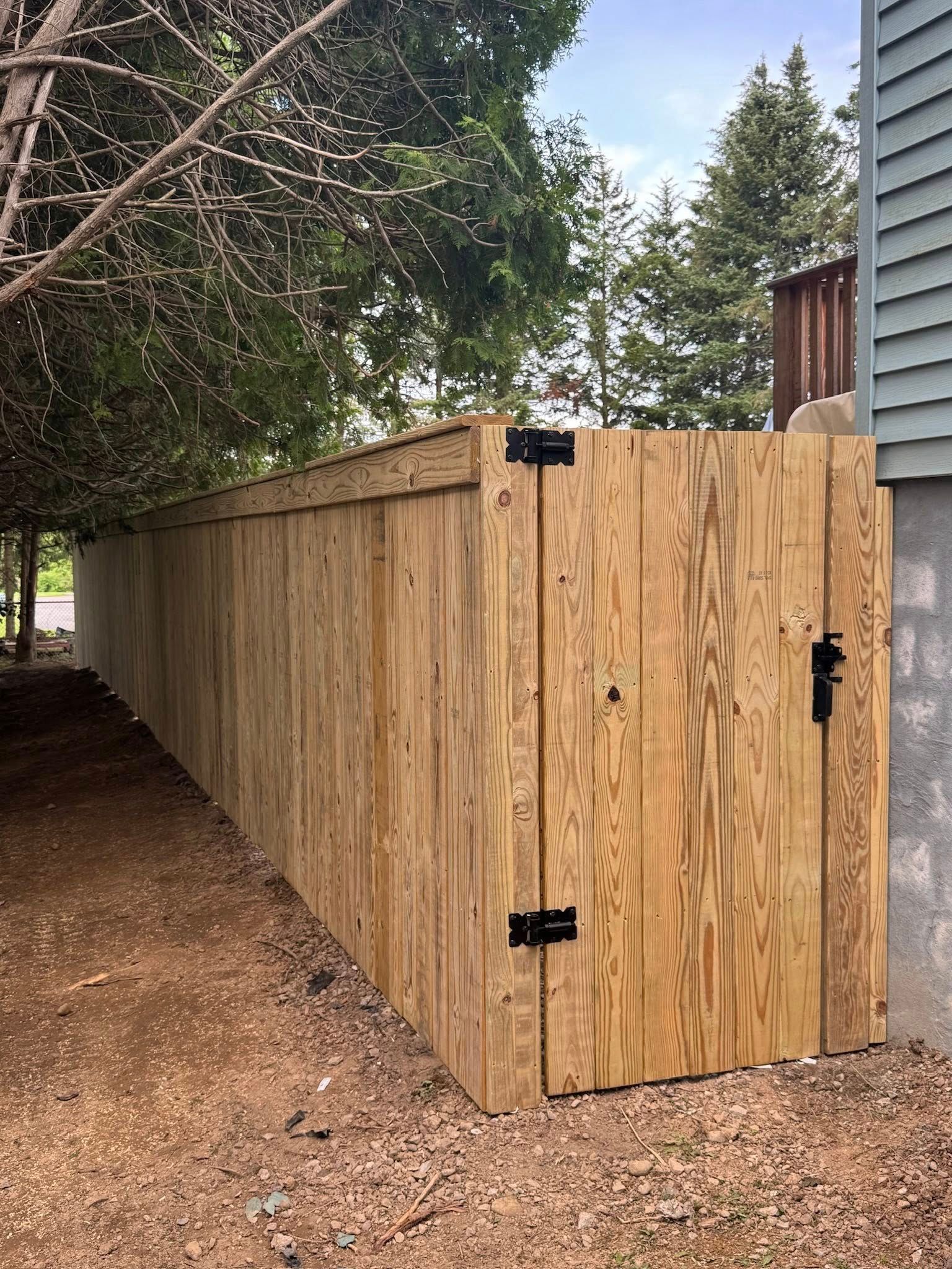 Wooden fence with a door, beside a building; gravel ground and overhanging trees.