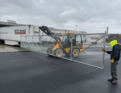 Yellow front-end loader lifts a chain-link fence. Worker in yellow hoody guides the fence near a loading dock.