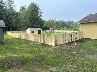 Wooden fence encloses a grassy yard with small sheds, surrounded by trees and a yellow house.
