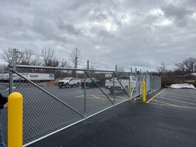 A chain-link fence with a sliding gate in a parking lot, with a white truck and yellow bollards.