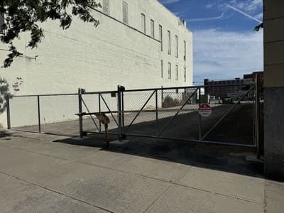 A metal gate secured by a chain-link fence blocks a driveway. The gate is in front of a large, white building.