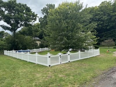 White picket fence surrounds a backyard pool and lawn, trees in the background, gray sky.
