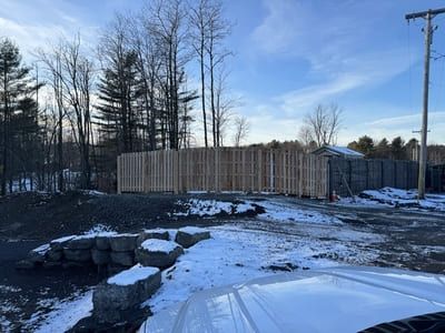 Wooden fence under construction next to a gravel lot, with snow on the ground and trees in the background.