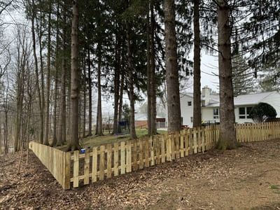Wooden picket fence along a wooded area, trees in the background, and a white house with a chimney.