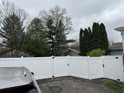 White vinyl fence in a driveway, trees in the background, overcast sky.