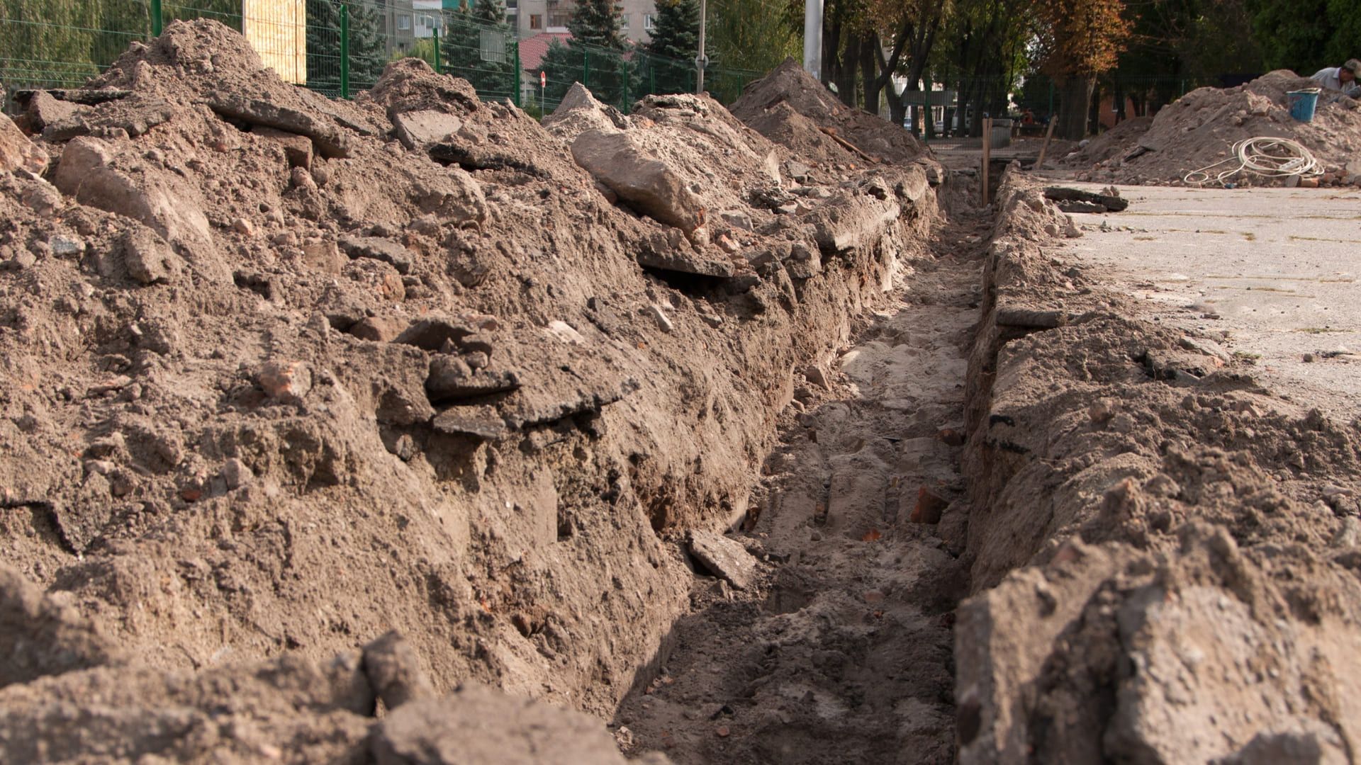 Long, narrow trench dug in dirt and asphalt, possibly for utilities, in an outdoor setting.