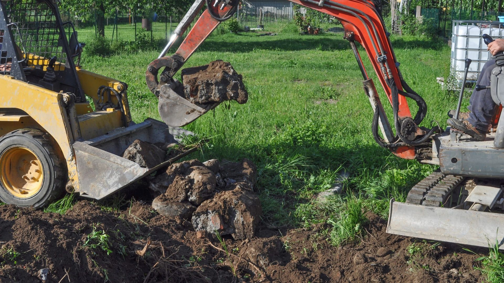 Mini excavator dumping rocks into the bucket of a yellow skid steer in a field.