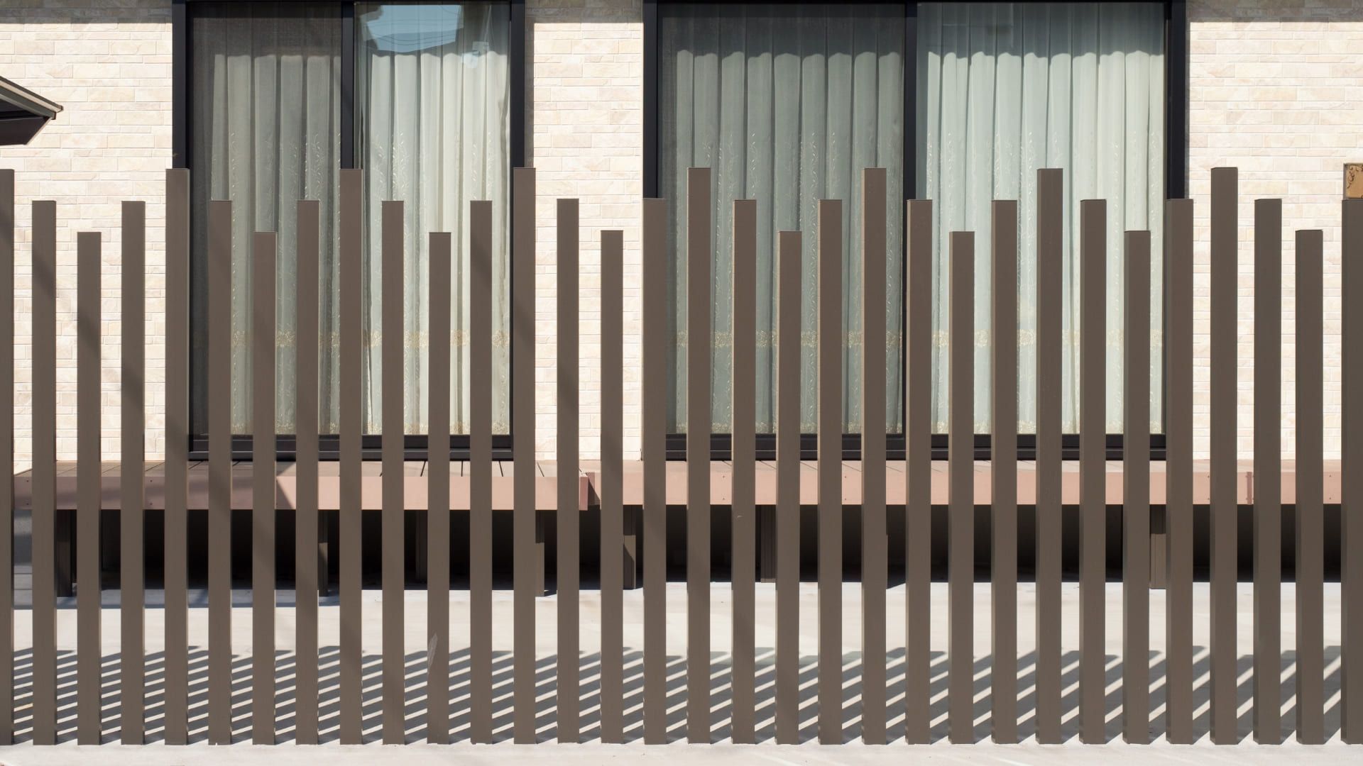 Brown vertical wooden fence in front of a building with windows. Shadows cast on the ground.